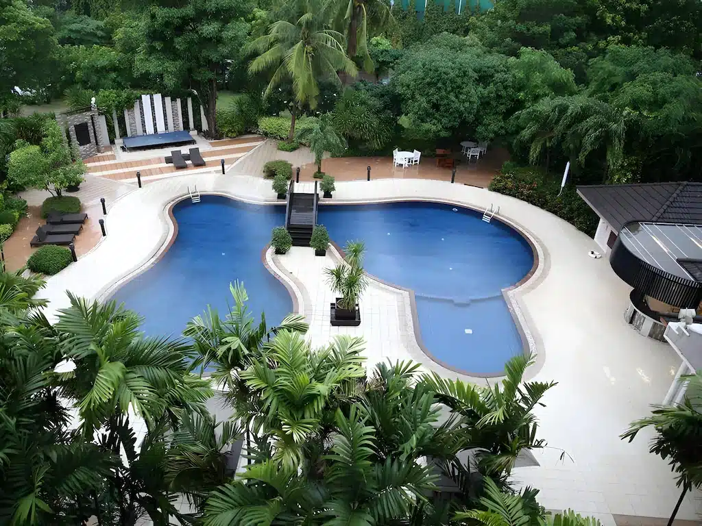 ภาพมุมสูงของสระว่ายน้ำกลางแจ้งขนาดใหญ่รูปทรงไม่สม่ำเสมอ ณ  โรงแรมกรุงเทพ โรงแรม 5 ดาวในกรุงเทพฯ ล้อมรอบด้วยต้นไม้ เก้าอี้อาบแดด และพื้นที่นั่งพักผ่อนในบรรยากาศสวนที่จัดแต่งอย่างสวยงาม.
