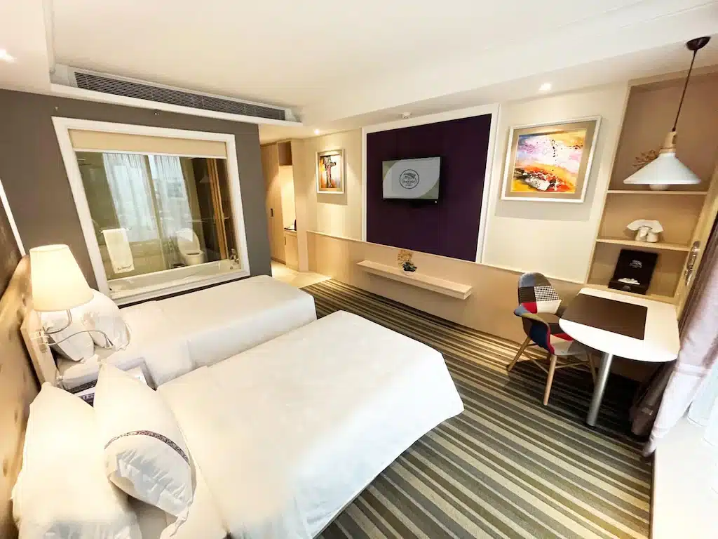 ห้องพักโรงแรมสมัยใหม่ที่ โรงแรมในกรุงเทพ พร้อมเตียงเดี่ยวสองเตียง โต๊ะทำงานและเก้าอี้ โทรทัศน์ติดผนัง พรมลายทาง งานศิลปะติดกรอบ และหน้าต่างกระจกบานใหญ่สู่ห้องน้ำ.