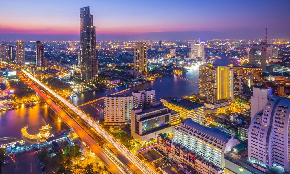 ภาพมุมสูงของทิวทัศน์เมืองยามพลบค่ำ พร้อมตึกสูงระฟ้า แม่น้ำที่ไหลผ่านกลางเมือง ถนนที่สว่างไสว และ โรงแรม 5 ดาวกรุงเทพ สุดหรูในกรุงเทพฯ ที่เพิ่มความสง่างามให้กับเส้นขอบฟ้าอันเปล่งประกาย.