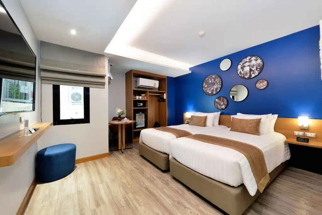 ห้องพักโรงแรมสมัยใหม่ในโรงแรมในกรุงเทพ ประกอบด้วยเตียงคู่สองเตียง ผนังสีน้ำเงินตัดกับกระจก โรงแรมในกรุงเทพ ตกแต่ง พื้นไม้ โต๊ะทำงาน โทรทัศน์ หน้าต่าง และเฟอร์นิเจอร์โทนสีเรียบเพื่อการพักผ่อนที่สะดวกสบาย.