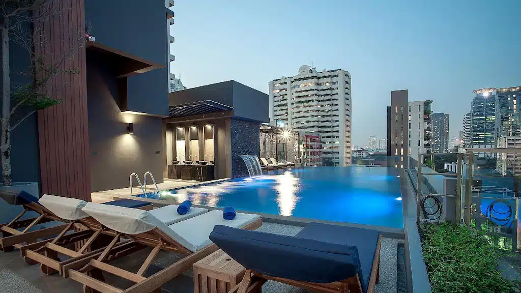 สระว่ายน้ำอินฟินิตี้บนดาดฟ้าพร้อม โรงแรมในกรุงเทพ เก้าอี้อาบแดดและทัศนียภาพของตึกสูงในเมืองยามพลบค่ำ มอบวิวเส้นขอบฟ้าของกรุงเทพฯ ที่สมบูรณ์แบบจากโรงแรมกรุงเทพแห่งนี้.
