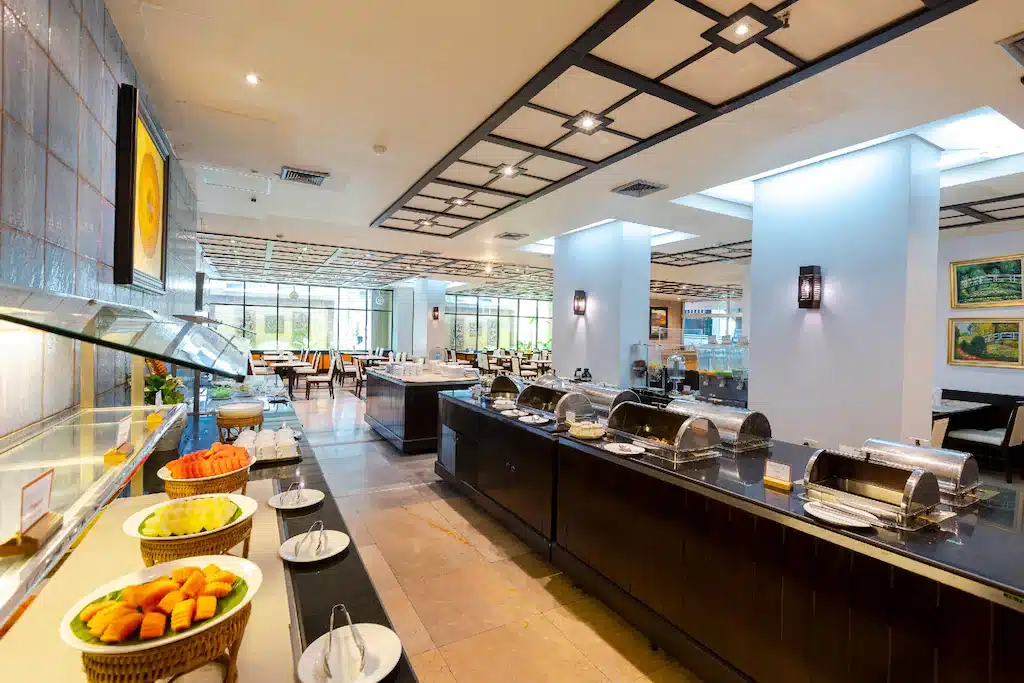 พื้นที่บุฟเฟ่ต์โรงแรมกว้างขวางที่โรงแรม 5 ดาวกรุงเทพ พร้อมถาดอาหารที่มี โรงแรมกรุงเทพ หลังคาคลุมบนเคาน์เตอร์, ผลไม้จัดเป็นถาดโชว์, และโต๊ะอาหารจัดเรียงไว้ด้านหลัง.