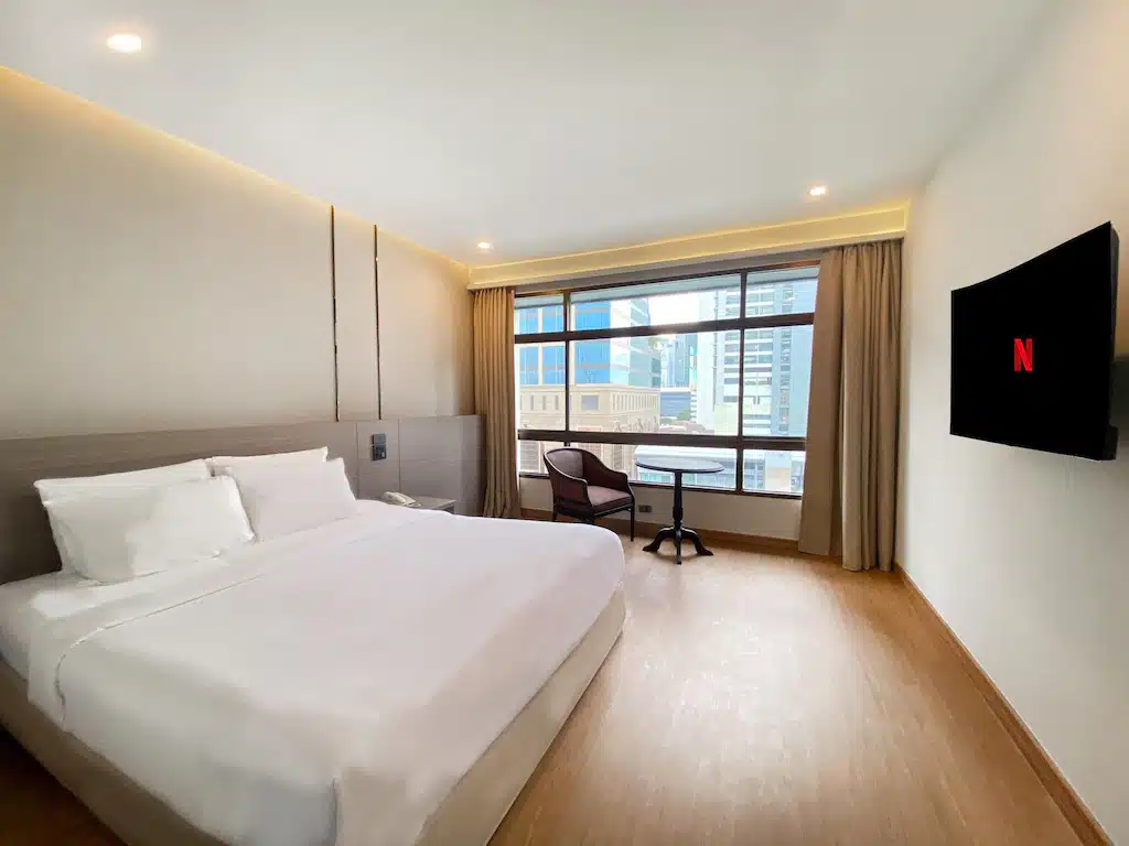 ห้องพักโรงแรมกรุงเทพฯ ทันสมัยพร้อมเตียงขนาดใหญ่ พื้นไม้ โต๊ะเล็กพร้อมเก้าอี้สองตัวริมหน้าต่าง และทีวีติดผนังแสดงโลโก้ Netflix. โรงแรมกรุงเทพ 
