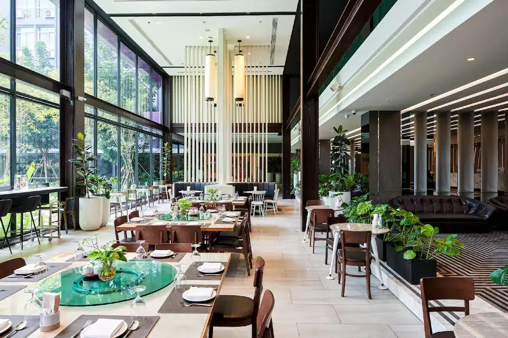 ร้านอาหารสมัยใหม่พร้อมหน้าต่างบานใหญ่จากพื้นจรดเพดาน โต๊ะและเก้าอี้ไม้ พืชสีเขียว และแสง โรงแรมกรุงเทพ ธรรมชาติที่ส่องเข้ามา—เหมาะสำหรับแขกที่ต้องการพื้นที่รับประทานอาหารที่สว่างไสวขณะเข้าพักที่ โรงแรมในกรุงเทพ.