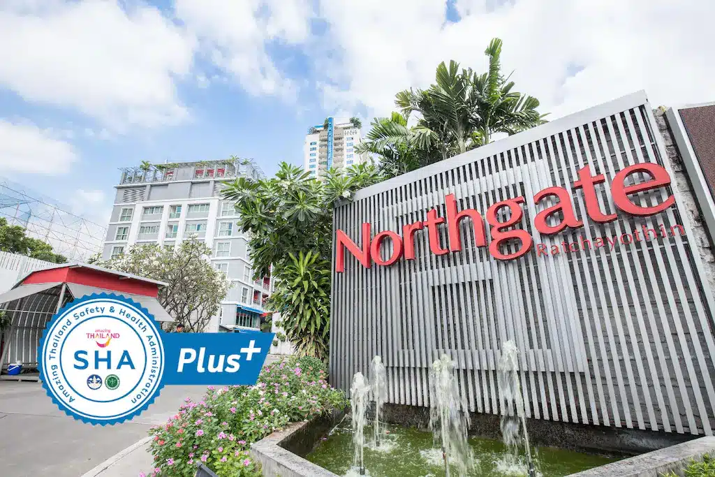มุมมองด้านหน้าของโรงแรม Northgate Ratchayothin ใน โรงแรมกรุงเทพ กรุงเทพฯ พร้อมป้ายสีแดงขนาดใหญ่ ต้นไม้เขียวชอุ่ม น้ำพุ และป้ายรับรอง SHA Plus+ อยู่ด้านหน้า เหมาะสำหรับผู้ที่มองหาที่พักระดับพรีเมียมในกรุงเทพฯ.