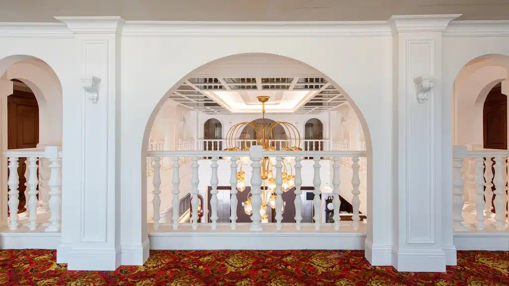 มุมมองผ่านซุ้มประตูสีขาวและราวบันไดไปยังโคมไฟระย้าอันวิจิตรที่แขวนอยู่ในห้องภายในที่กว้างขวางและหรูหรา พร้อมพรมลายสีแดงที่ โรงแรมในกรุงเทพ