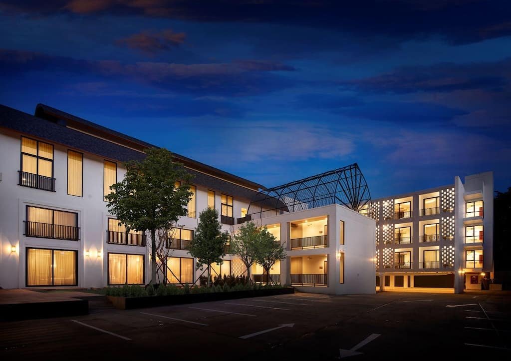 โรงแรมสมัยใหม่สูงสามชั้น โดดเด่นด้วยหน้าต่างที่ส่องแสงสว่าง ระเบียง และลานจอดรถที่เกือบว่างเปล่าในยามพลบค่ำ ท่ามกลางท้องฟ้ายามเย็นสีคราม—ที่พักอันน่าดึงดูดสำหรับผู้ที่กำลังสำรวจสถานที่ท่องเที่ยวในน่าน.