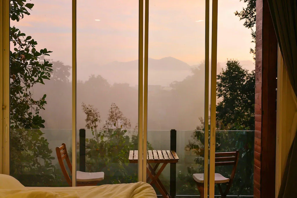 โต๊ะไม้ขนาดเล็กพร้อมเก้าอี้สองตัวตั้งอยู่บนระเบียงที่มองเห็นต้นไม้และภูเขาที่ปกคลุมด้วยหมอก ผ่านประตูกระจกยามรุ่งอรุณ—เหมาะสำหรับผู้ที่มองหาสถานที่ท่องเที่ยวในน่านและเสน่ห์อันเงียบสงบของการท่องเที่ยวในน่านในช่วงเช้าตรู่.