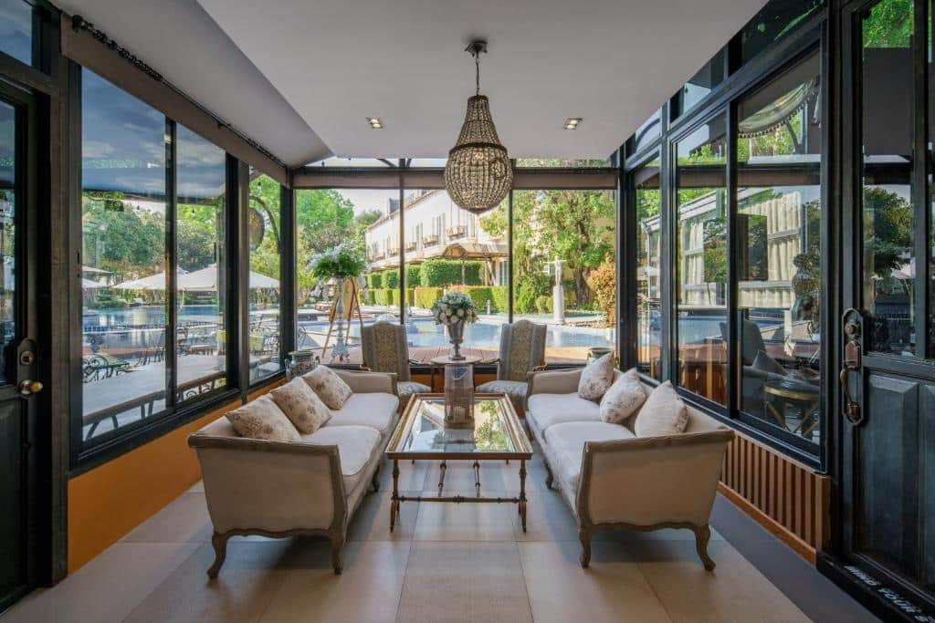 พื้นที่เลานจ์ที่ล้อมรอบด้วยกระจกพร้อมโซฟาสองตัว เก้าอี้นวม โต๊ะกาแฟ และโคมไฟระย้า มองเห็นสระว่ายน้ำกลางแจ้งและสวน—เหมาะสำหรับการพักผ่อนหลังจากสำรวจแหล่งท่องเที่ยวเขาใหญ่.