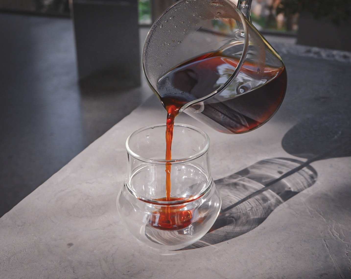 เหยือกแก้วรินกาแฟลงในถ้วยแก้วสองชั้นบนพื้นผิวสีอ่อน แสงแดดส่องทอดเงา—เหมาะสำหรับการผ่อนคลายหลังจากสำรวจสถานที่ท่องเที่ยวเขาใหญ่.