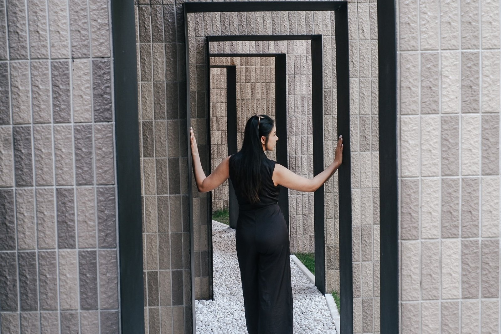 ผู้หญิงในชุดสีดำยืนอยู่ระหว่างกรอบโลหะรูปสี่เหลี่ยมผืนผ้ากลางแจ้งที่ ที่เที่ยวเขาใหญ่ สัมผัสกับด้านข้าง ล้อมรอบด้วยกำแพงที่มีพื้นผิวสีเบจและกรวดสีขาว.