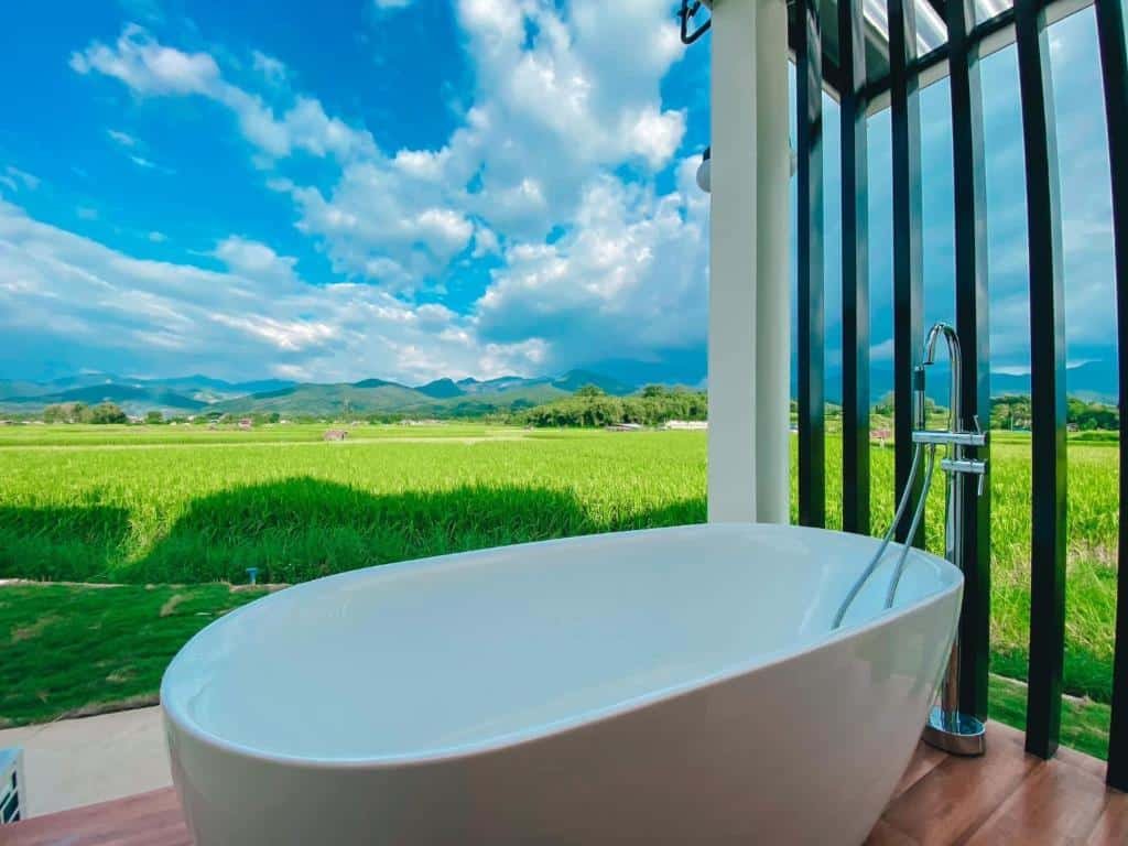 อ่างอาบน้ำสีขาวตั้งอยู่บนระเบียงไม้พร้อมวิวทุ่งหญ้าสีเขียว ภูเขาไกล และท้องฟ้าที่มีเมฆบางส่วน—เหมาะสำหรับการผ่อนคลายขณะสำรวจสถานที่ท่องเที่ยวในน่าน.