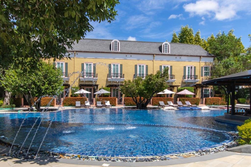 สระว่ายน้ำกลางแจ้งขนาดใหญ่พร้อมน้ำพุ เก้าอี้อาบแดด ร่มไม้ ต้นไม้ และตึกสองชั้นสีเหลืองเป็นฉากหลังภายใต้ท้องฟ้าสีคราม—เหมาะสำหรับการผ่อนคลายที่ สถานที่ท่องเที่ยวเขาใหญ่ ชั้นยอด.