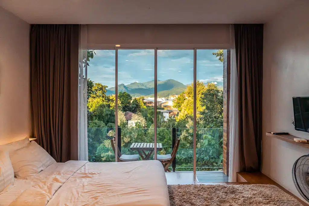 ห้องพักโรงแรมพร้อมเตียงคู่ ประตูกระจกเปิดออกสู่ระเบียงที่มีโต๊ะและเก้าอี้สองตัว พร้อมวิวภูเขา—เหมาะสำหรับการพักผ่อนหลังจากสำรวจสถานที่ท่องเที่ยวในน่านและสถานที่ท่องเที่ยวที่สวยงามในน่าน.