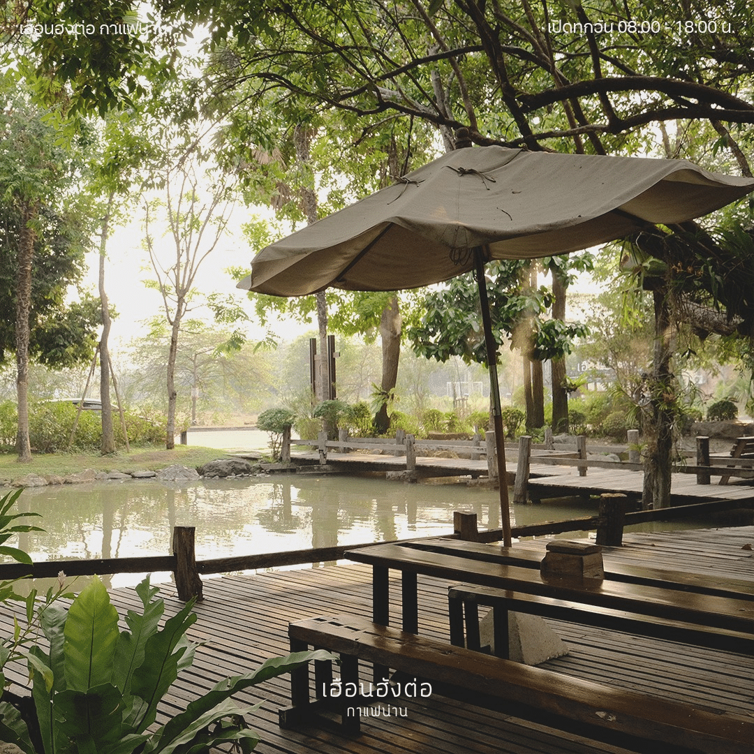 พื้นที่นั่งเล่นกลางแจ้งพร้อมโต๊ะและม้านั่งไม้ ริมสระน้ำ ใต้ร่มเงาไม้และร่มสีเบจขนาดใหญ่—เหมาะสำหรับการพักผ่อนหรือพบปะสังสรรค์กับเพื่อนฝูง ท่ามกลางบรรยากาศธรรมชาติ จุดเช็คอินยอดนิยมของสถานที่ท่องเที่ยวในน่าน.