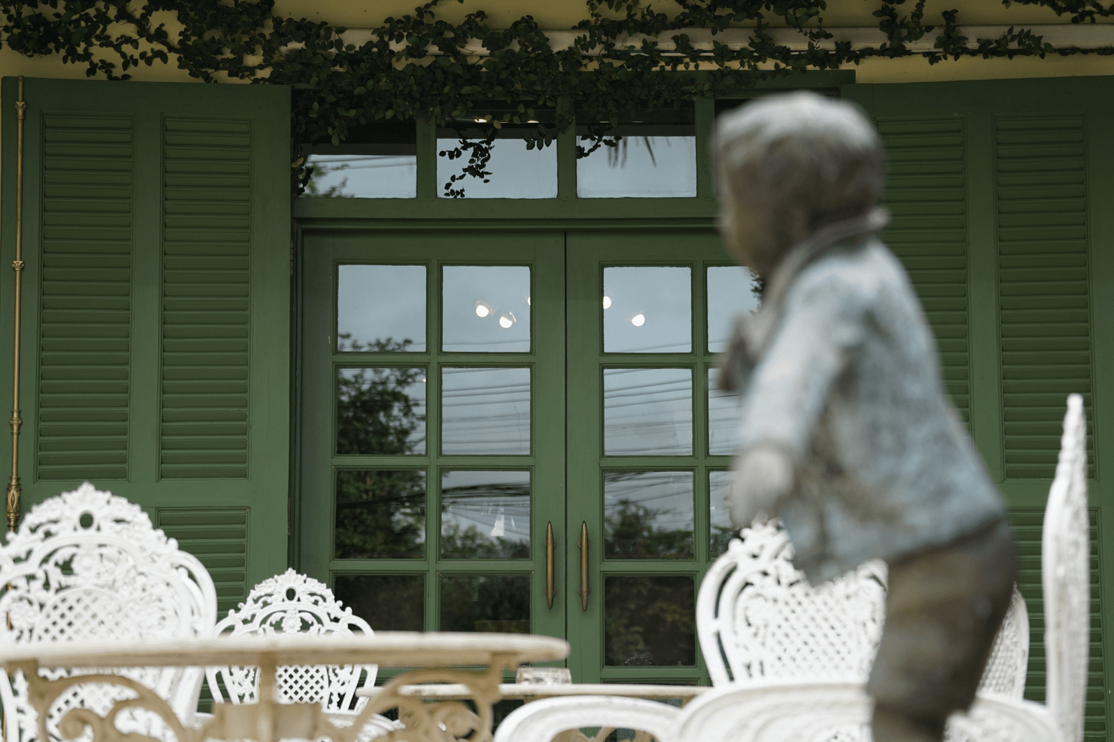 ประตูฝรั่งเศสสีเขียวพร้อมบานหน้าต่างที่เข้าชุดกันและเฟอร์นิเจอร์ลานระเบียงสีขาวประดับลวดลายวิจิตรในเบื้องหน้า สร้างเสน่ห์ของ น่านที่เที่ยว ในขณะที่รูปปั้นเด็กที่เบลอเพิ่มความงดงามทางศิลปะ.