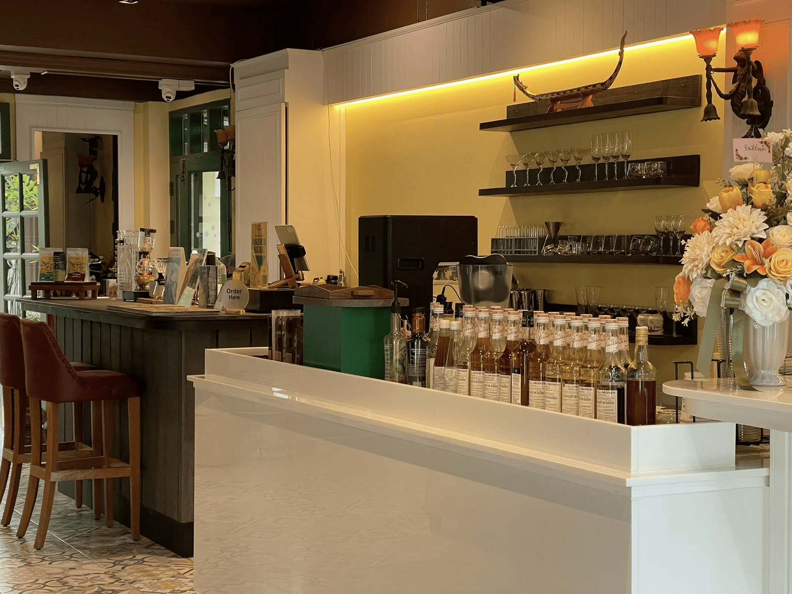 เคาน์เตอร์คาเฟ่แสนอบอุ่นใน น่านที่เที่ยว พร้อมขวดน้ำเชื่อม แก้วบนชั้นวาง เครื่องคิดเงิน ช่อดอกไม้สด และเก้าอี้บาร์ริมบาร์ไม้—เหมาะสำหรับการพักผ่อนขณะที่คุณเที่ยว น่าน.