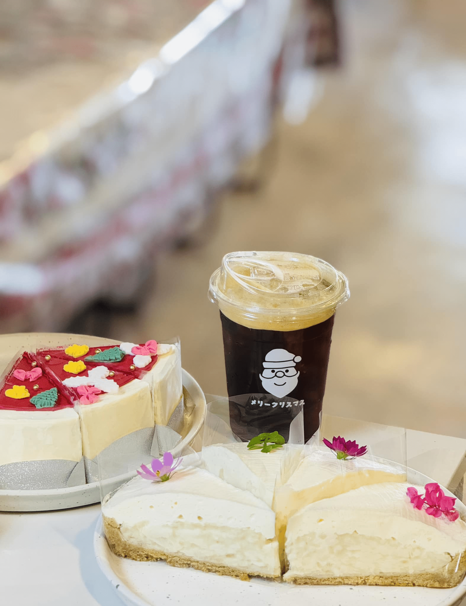 เค้กชีสเค้กสองจานพร้อมดอกไม้ตกแต่งและแก้วกาแฟเย็นที่มีโลโก้การ์ตูนอยู่บนโต๊ะ เหมาะสำหรับการผ่อนคลายที่คาเฟ่สุดชิคใกล้สถานที่ท่องเที่ยวในน่าน.
