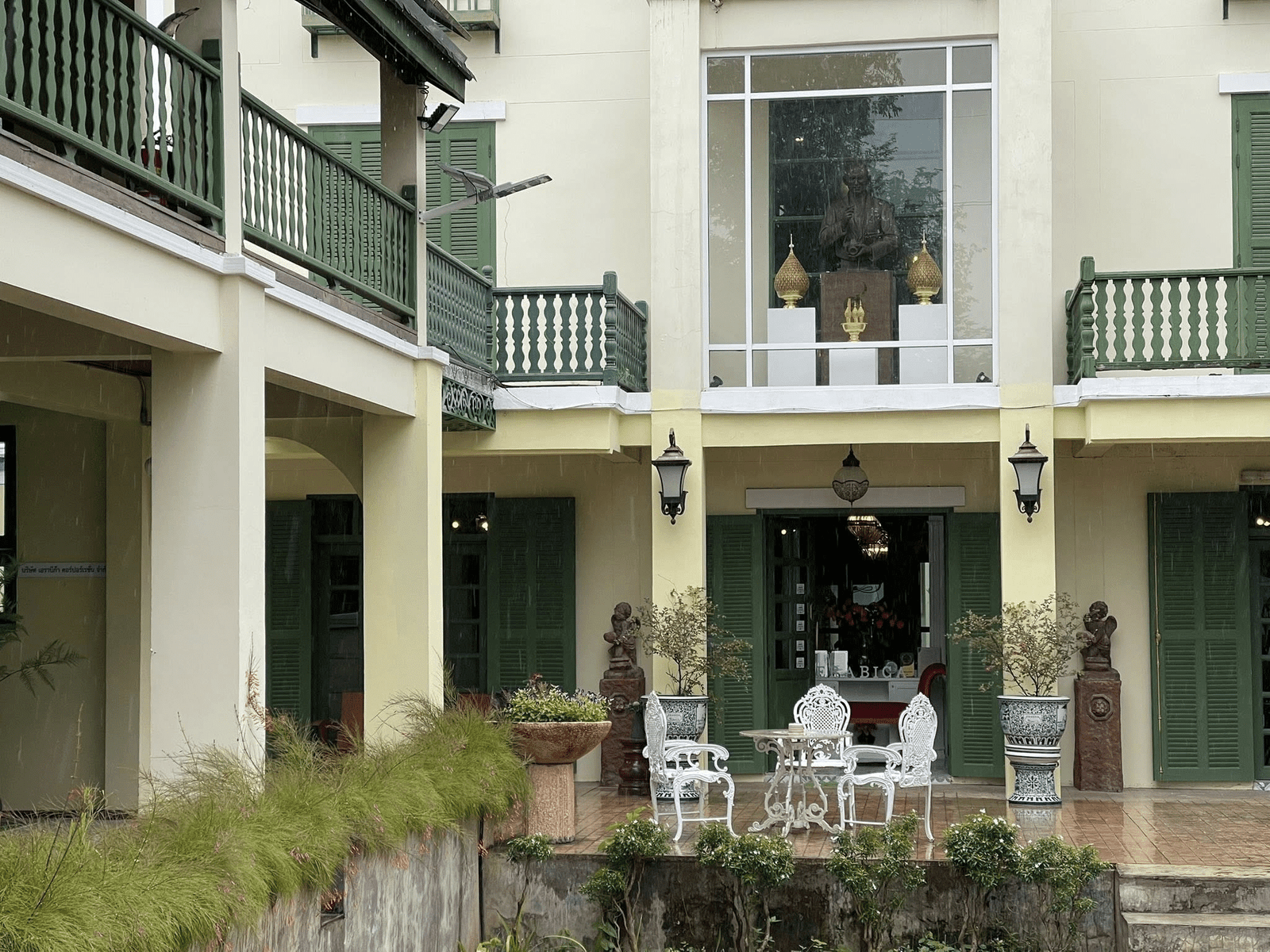 โต๊ะโลหะสีขาวพร้อมเก้าอี้สองตัวตั้งอยู่บนลานระเบียงหน้าตึกสีครีมที่มีบานหน้าต่างและระเบียงสีเขียว สร้างบรรยากาศน่ารักเหมาะสำหรับการพักผ่อนระหว่างการผจญภัยที่ น่าน.