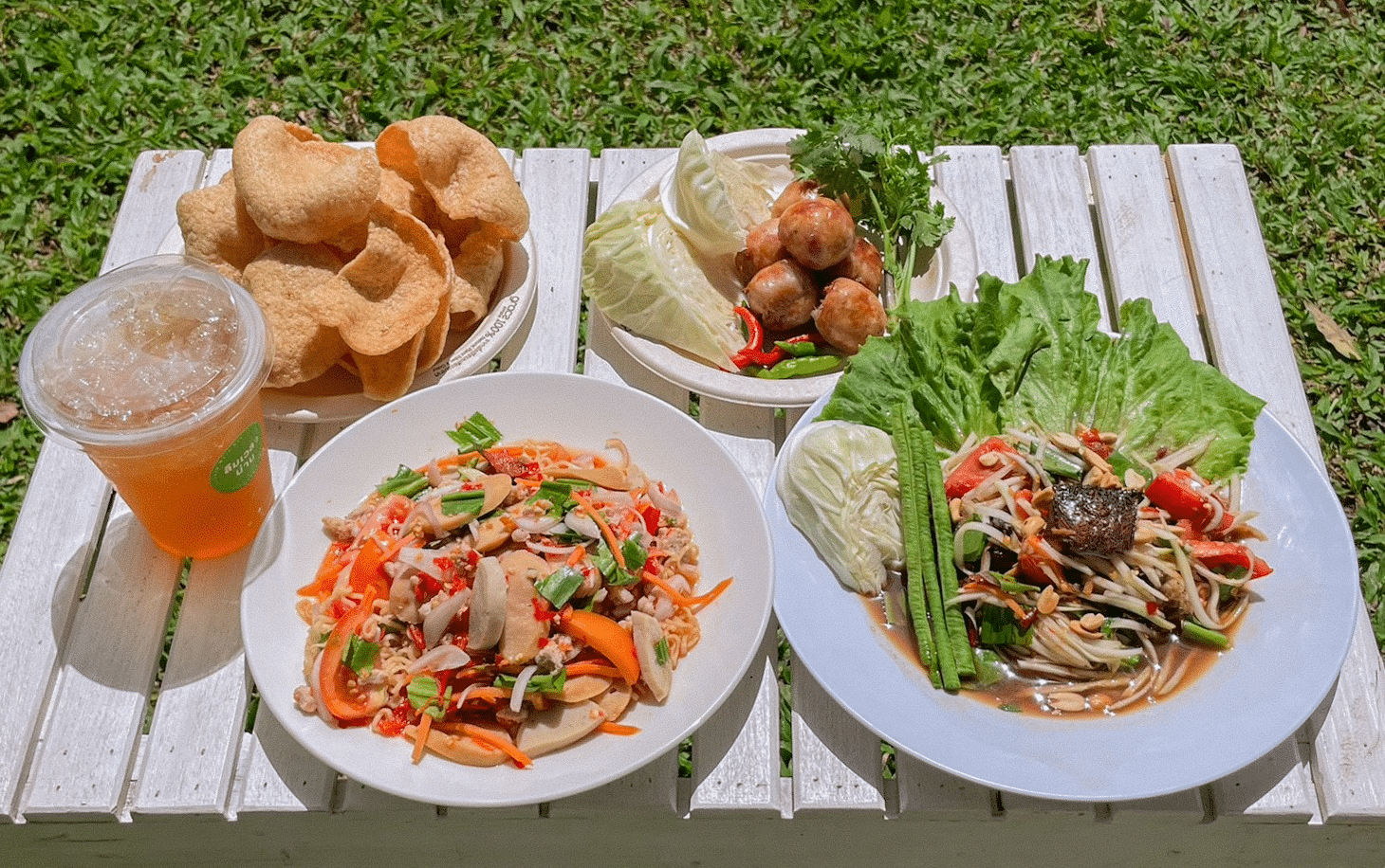 โต๊ะสีขาวที่จัดวางพร้อมชาเย็น, ชิปกุ้ง, ไส้กรอกกับผัก, และจานสลัดไทยสองจาน ตั้งอยู่นอกบ้านบนสนามหญ้า—เหมาะสำหรับการเพลิดเพลินกับรสชาติที่เป็นเอกลักษณ์ในขณะที่คุณสำรวจสถานที่ท่องเที่ยวในน่าน.