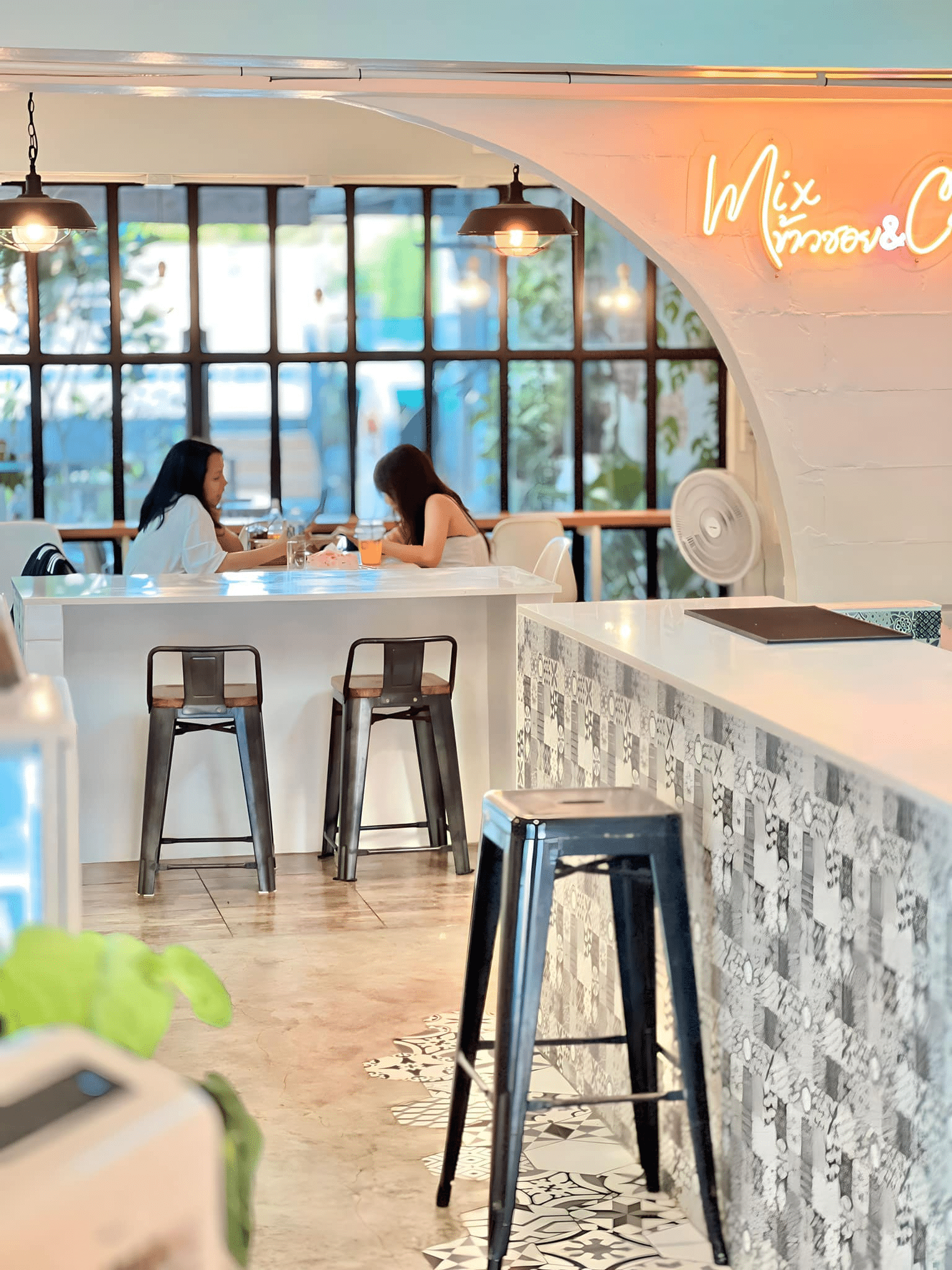 ผู้หญิงสองคนนั่งอยู่ที่โต๊ะคุยกันในคาเฟ่สมัยใหม่ที่มีบาร์กระเบื้องลาย, เก้าอี้สูง, หน้าต่างบานใหญ่, และป้ายนีออน—สถานที่อบอุ่นเหมาะสำหรับการเที่ยวเมืองน่านหรือค้นหาสถานที่ท่องเที่ยวใหม่ ๆ ในเมืองน่าน.