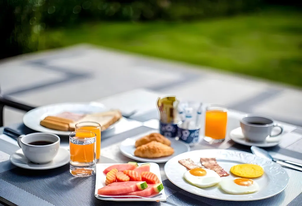 โต๊ะอาหารเช้าที่จัดเตรียมไว้พร้อมไข่, เบคอน, ขนมปังปิ้ง, ครัวซองต์, ผลไม้หั่น, น้ำส้ม, และกาแฟกลางแจ้งในวันที่แดดสดใส—เหมาะอย่างยิ่งสำหรับการเริ่มต้นวันใหม่ก่อนออกไปสำรวจสถานที่ท่องเที่ยวที่นิยมในน่าน.