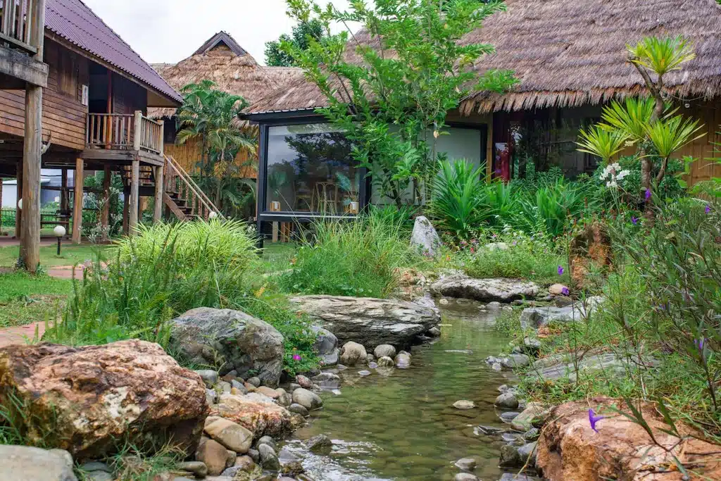 ลำธารเล็กๆ ไหลผ่านสวนที่จัดแต่งอย่างสวยงาม ล้อมรอบด้วยอาคารไม้และหลังคามุงจาก พร้อมพืชสีเขียวชอุ่มและก้อนหิน—สถานที่เงียบสงบสำหรับผู้ที่มองหา เที่ยวเมืองน่าน และสำรวจสถานที่ท่องเที่ยวที่ไม่เหมือนใครใน น่าน.