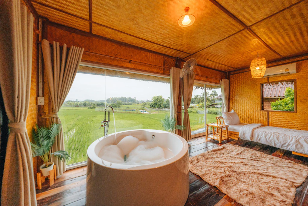 อ่างอาบน้ำทรงกลมที่เต็มไปด้วยฟองสบู่ตั้งอยู่ในห้องไม้ที่มีหน้าต่างบานใหญ่เปิดรับวิวทุ่งหญ้าสีเขียว—สถานที่หลบหนีอันน่าดึงดูดสำหรับผู้ที่มองหา น่านที่เที่ยว พร้อมด้วยเตียงนอนแสนสบาย พรมปูพื้น และต้นไม้กระถางเขียวชอุ่ม.