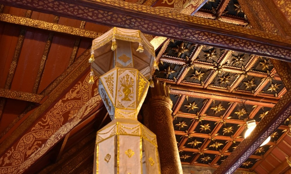 เพดานและเสาไม้ที่ตกแต่งอย่างประณีตด้วยลวดลายดอกไม้สีทองและรูปทรงเรขาคณิตในอาคารแบบดั้งเดิม เหมาะสำหรับผู้ที่มองหาสถานที่ท่องเที่ยวในน่านเพื่อชื่นชมงานฝีมืออันวิจิตร.