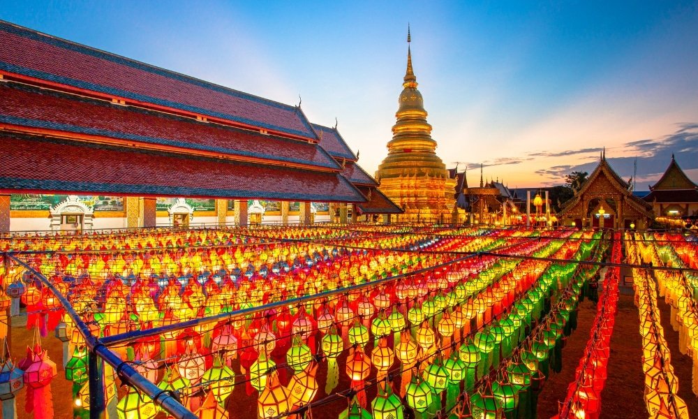 แถวโคมไฟหลากสีสันส่องสว่างยามพระอาทิตย์ตกดินเบื้องหน้าเจดีย์สีทอง แสดงให้เห็นถึงสถาปัตยกรรมไทยแบบดั้งเดิม—เป็นภาพที่น่าตื่นตาตื่นใจและต้องไปชมให้ได้สำหรับใครก็ตามที่กำลังสำรวจสถานที่ท่องเที่ยวในน่าน.