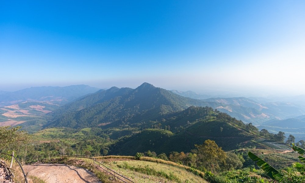 เทือกเขาที่มีเนินเขาเขียวขจีและต้นไม้ภายใต้ท้องฟ้าสีฟ้าใส มองเห็นจากจุดชมวิวสูงในวันที่แดดจ้า—เหมาะสำหรับผู้ที่กำลังมองหาสถานที่ท่องเที่ยวในน่านหรือสถานที่ท่องเที่ยวใหม่ในน่านเพื่อสำรวจ.