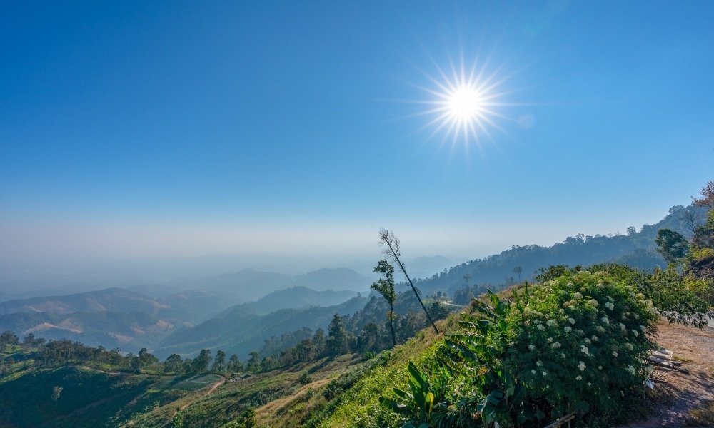 ท้องฟ้าแจ่มใสและสดใสเหนือเนินเขาสีเขียวและภูเขาไกลโพ้นใน น่านที่เที่ยว พร้อมด้วยพืชพรรณหนาแน่นในเบื้องหน้า และดวงอาทิตย์ส่องแสงสว่างไสวเหนือขอบฟ้า.