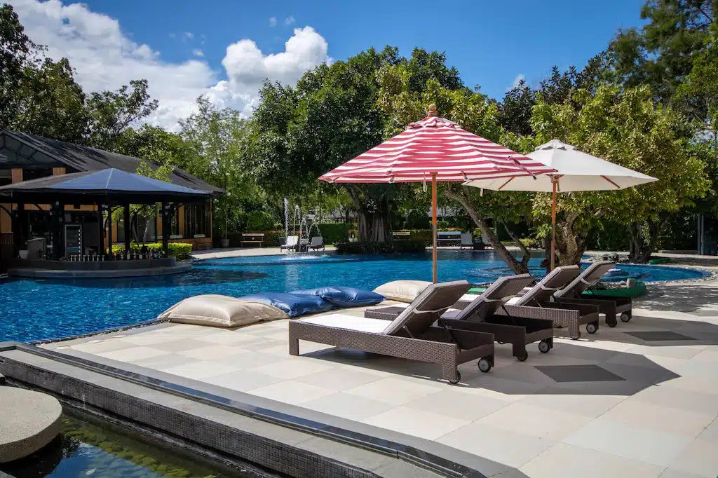พื้นที่สระว่ายน้ำกลางแจ้งพร้อมเก้าอี้หวายสำหรับนั่งพักผ่อน ร่มกันแดด และเบาะรองนั่งขนาดใหญ่ริมสระว่ายน้ำสีฟ้าใสสะอาด ล้อมรอบด้วยต้นไม้และอาคาร—เหมาะอย่างยิ่งสำหรับการพักผ่อนระหว่างการเดินทางท่องเที่ยวเขาใหญ่ของคุณ.