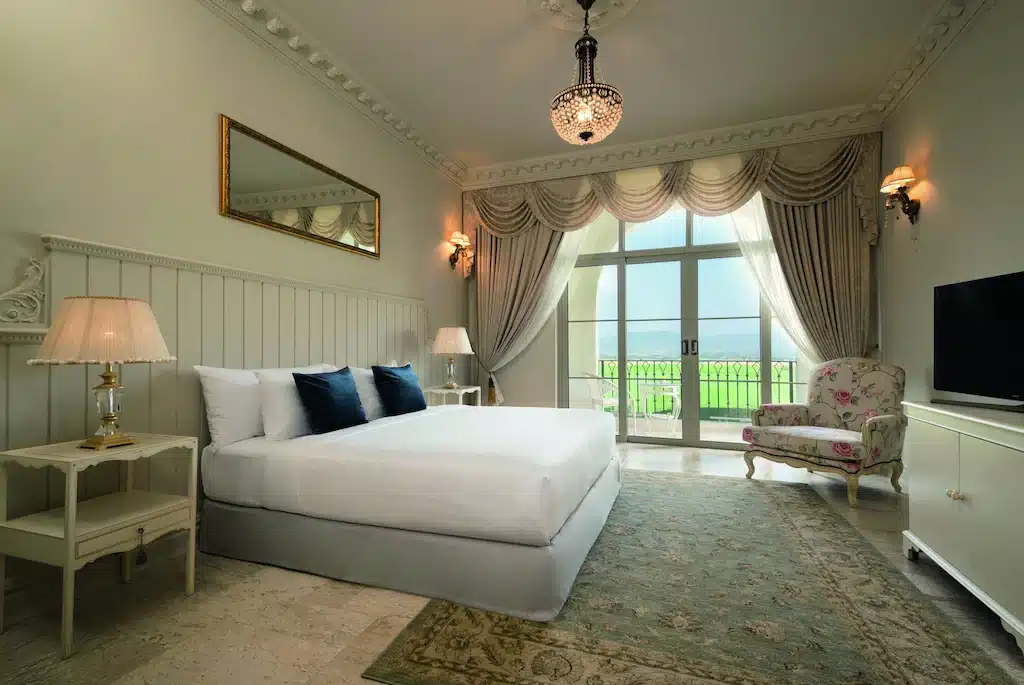 ห้องนอนหรูหราพร้อมเตียงขนาดใหญ่ โต๊ะข้างเตียง เก้าอี้นวม โทรทัศน์ และพรม ประตูฝรั่งเศสเปิดออกสู่ระเบียงที่มีวิวทิวทัศน์สีเขียว—เหมาะสำหรับการพักผ่อนหลังจากสำรวจแหล่งท่องเที่ยวเขาใหญ่.