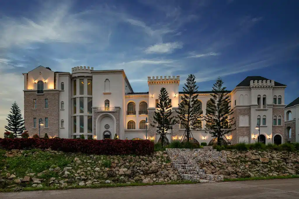 อาคารขนาดใหญ่หลายชั้นที่มีลักษณะคล้ายปราสาท ส่องแสงสว่างในยามพลบค่ำ ล้อมรอบด้วยต้นสนและบันไดหินที่นำไปสู่ทางเข้า—สถานที่ที่น่าหลงใหลสำหรับผู้ที่กำลังมองหา ที่เที่ยวเขาใหญ่.