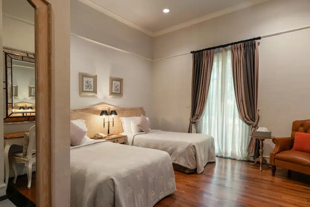 ห้องพักโรงแรมพร้อมเตียงเดี่ยวสองเตียง โต๊ะข้างเตียง เก้าอี้นวม หน้าต่างที่มีผ้าม่านติดไว้ กระจกติดผนัง และภาพติดกรอบเหนือเตียง—เหมาะสำหรับการผ่อนคลายหลังจากเที่ยวชมสถานที่ท่องเที่ยวเขาใหญ่.