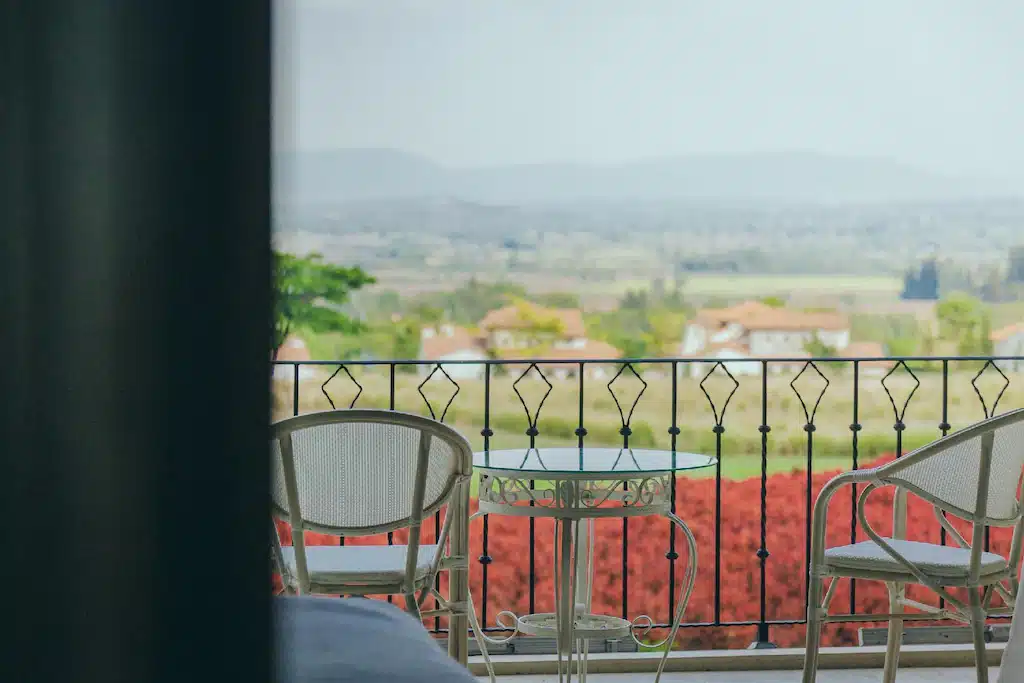 โต๊ะและเก้าอี้โลหะสองตัวบนระเบียงมองเห็นทิวทัศน์ชนบทที่สวยงามพร้อมต้นไม้สีแดงและภูเขาไกล ๆ—เหมาะสำหรับการผ่อนคลายหลังจากวันที่ออกไปเที่ยวเขาใหญ่.