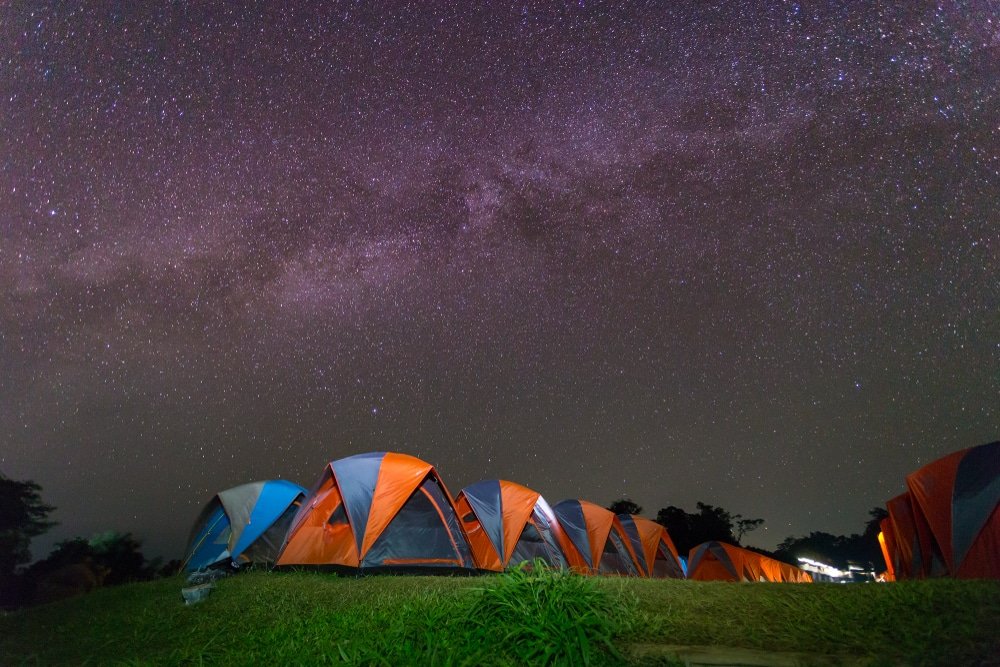 แถวเต็นท์สีสันสดใสตั้งเรียงรายบนสนามหญ้าใต้ท้องฟ้ายามค่ำคืนที่แจ่มใสเต็มไปด้วยดวงดาวและทางช้างเผือก—เหมาะสำหรับการสัมผัสธรรมชาติที่ น่านที่เที่ยว.