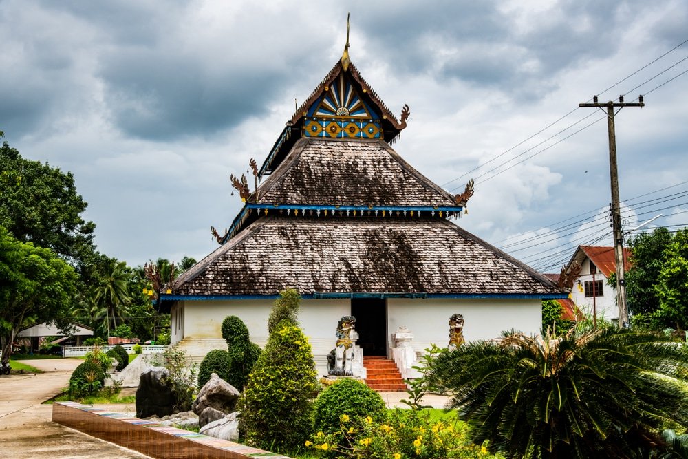 วัดแบบดั้งเดิมที่มีหลังคาสูงชันหลายชั้น ประดับด้วยรูปปั้นและต้นไม้สีเขียว ตั้งอยู่ใต้ท้องฟ้าที่มีเมฆครึ้ม โดยมีสายไฟอยู่ทางขวา—สถานที่ท่องเที่ยวที่เงียบสงบในน่านสำหรับนักท่องเที่ยวที่จะมาสำรวจ.