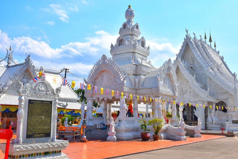 วัดสีขาวที่มีลวดลายแกะสลักและตกแต่งอย่างประณีต ล้อมรอบด้วยธงสีสันสดใสและแผ่นป้ายหน้าวัดใต้ท้องฟ้าที่มีเมฆบางส่วน—สถานที่ท่องเที่ยวที่ไม่ควรพลาดในน่านสำหรับผู้ที่มาเยือนภูมิภาคที่มีเสน่ห์แห่งนี้.