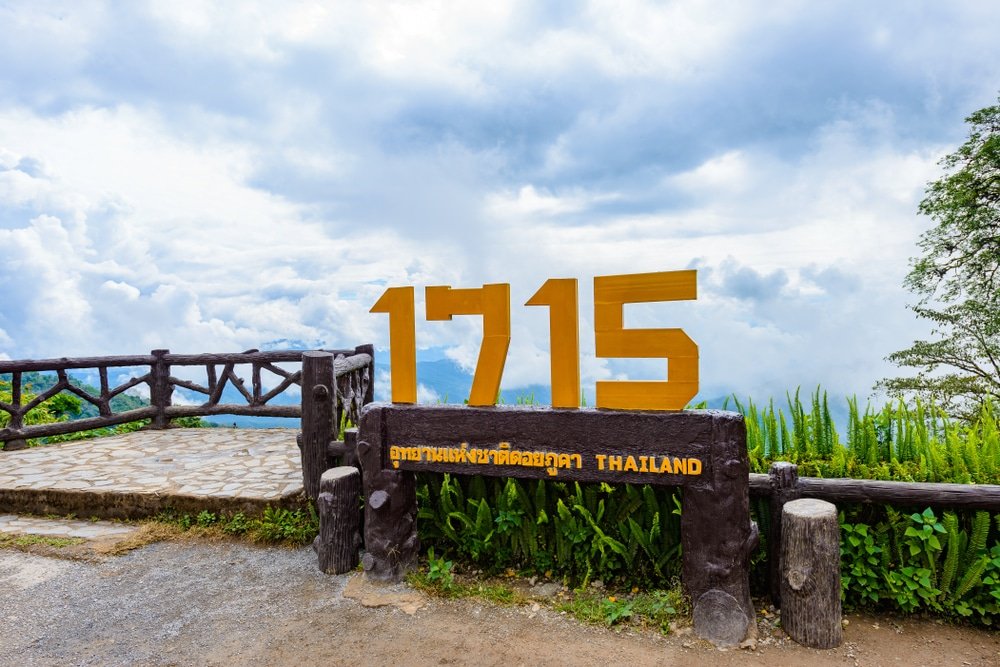 ป้ายสีเหลือง "1715" พร้อมคำว่า "ประเทศไทย" ด้านล่าง ระบุจุดชมวิวที่รายล้อมไปด้วยต้นไม้เขียวขจีและท้องฟ้าที่มีเมฆปกคลุม—จุดที่เป็นสัญลักษณ์สำหรับผู้ที่มาเยือน เที่ยวน่าน.