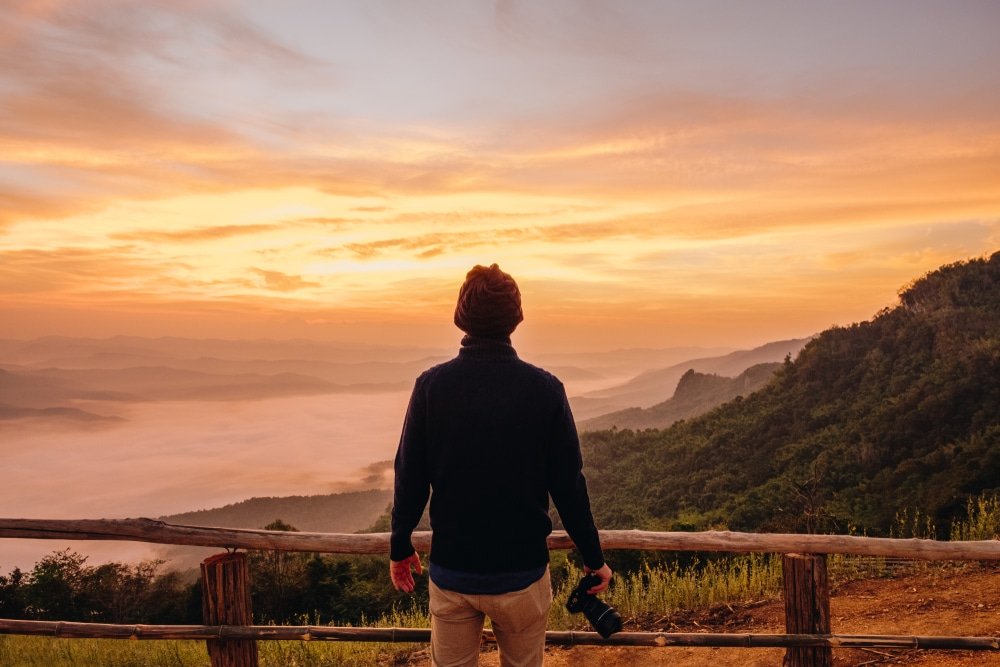 บุคคลหนึ่งยืนอยู่บนแท่นไม้ที่มองเห็นหุบเขาและภูเขาที่ปกคลุมด้วยหมอกยามพระอาทิตย์ขึ้น มือข้างหนึ่งถือกล้องถ่ายรูปไว้—เหมาะอย่างยิ่งสำหรับการบันทึกความงดงามอันน่าทึ่งของสถานที่ท่องเที่ยวในน่าน.