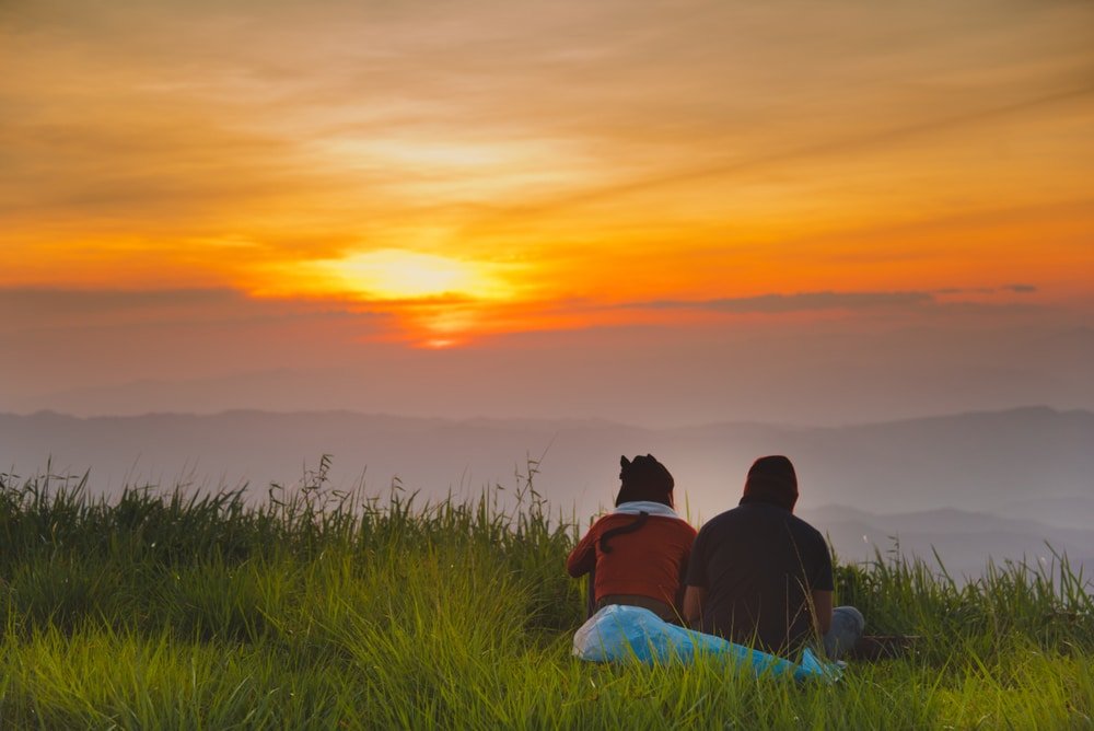 สองคนนั่งอยู่บนพื้นหญ้า หน้ามองพระอาทิตย์ตกสีส้มและเหลืองที่สดใสเหนือภูเขาไกลและทิวทัศน์ที่มีหมอกปกคลุม โอบกอดความสวยงามที่สงบของ เที่ยวน่าน.