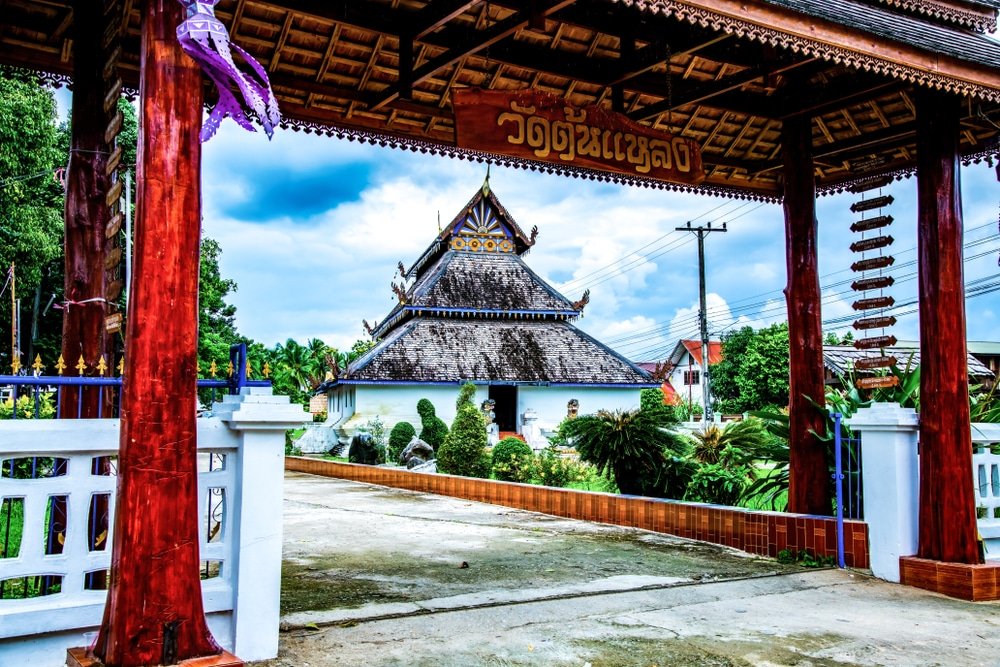 วัดไทยแบบดั้งเดิมที่มีหลังคาหลายชั้นมองเห็นได้ผ่านประตูไม้ล้อมรอบด้วยต้นไม้เขียวขจีและท้องฟ้าที่มีเมฆครึ้ม—สถานที่ท่องเที่ยวที่ไม่ควรพลาดในน่านสำหรับผู้ที่ต้องการสำรวจความงามของเมืองน่าน.