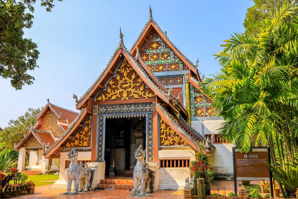 วัดไทยแบบดั้งเดิมที่มีหน้าไม้แกะสลักอย่างประณีต ประดับด้วยทองคำอย่างละเอียด และมีรูปปั้นผู้พิทักษ์สองตนที่ทางเข้า ล้อมรอบด้วยต้นไม้เขียวชอุ่ม—สถานที่ท่องเที่ยวที่ไม่ควรพลาดในน่านสำหรับผู้ที่ต้องการเที่ยวชมเมืองน่าน.