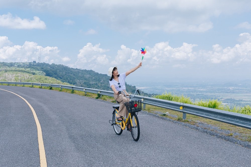 คนขี่จักรยานสีเหลืองบนถนนคดเคี้ยวที่ ที่เที่ยวเขาใหญ่, ถือกังหันสีสันสดใสด้วยมือข้างหนึ่ง ภูเขา, หญ้าเขียว, และเมฆสร้างฉากหลังที่สมบูรณ์แบบสำหรับการผจญภัย เที่ยวเขาใหญ่ นี้.