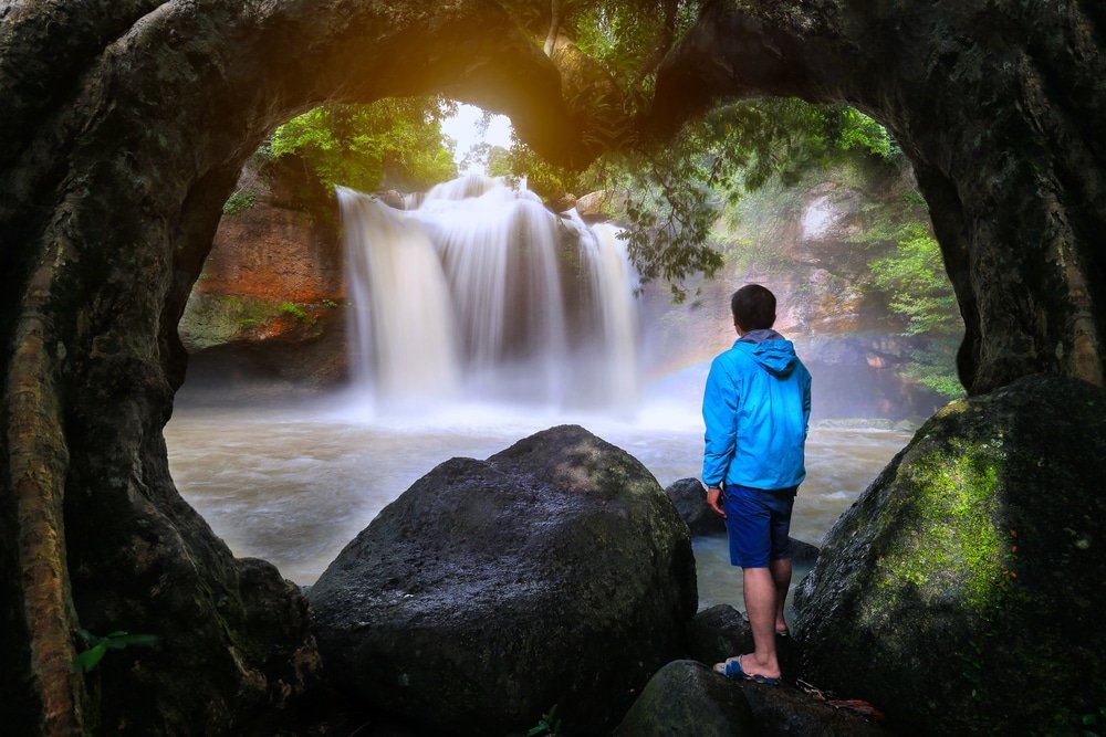 บุคคลในเสื้อแจ็คเก็ตสีน้ำเงินยืนอยู่บนโขดหิน มองดูน้ำตกที่ถูกล้อมรอบด้วยกิ่งไม้และพืชพรรณเขียวขจี—ช่วงเวลาอันเงียบสงบในการสำรวจสถานที่ท่องเที่ยวเขาใหญ่.