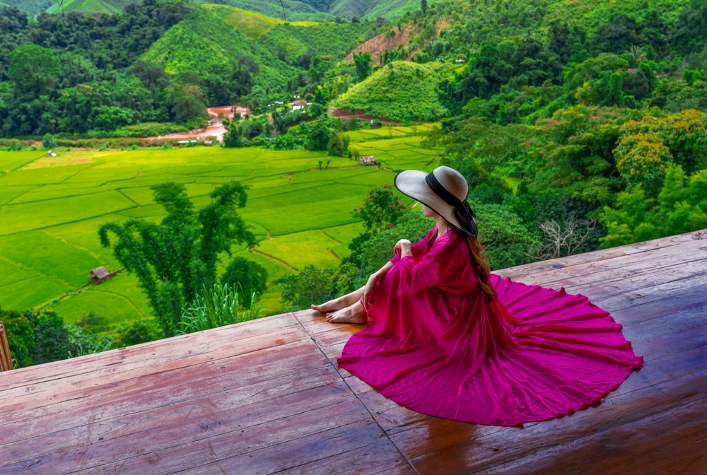 บุคคลในหมวกปีกกว้างและชุดกระโปรงสีแดงพลิ้ว นั่งอยู่บนแท่นไม้ ดื่มด่ำกับความงดงามของเนินเขาสีเขียวและทุ่งนาข้าว—เหมาะอย่างยิ่งสำหรับผู้ที่กำลังมองห สถานที่ท่องเที่ยวในน่าน.