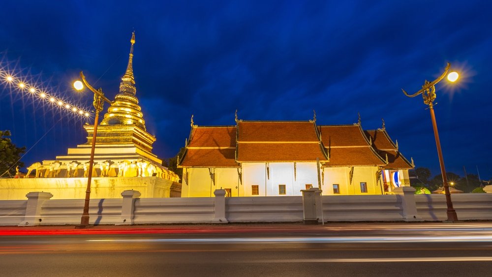 พระธาตุทองคำและวัดแบบดั้งเดิมส่องแสงสว่างในยามค่ำคืนที่เมืองน่าน หนึ่งในสถานที่ท่องเที่ยวที่ไม่ควรพลาดของน่าน โดยมีแสงไฟจากรถยนต์ที่ผ่านไปมาบนถนนด้านหน้าเป็นภาพเบลอ.