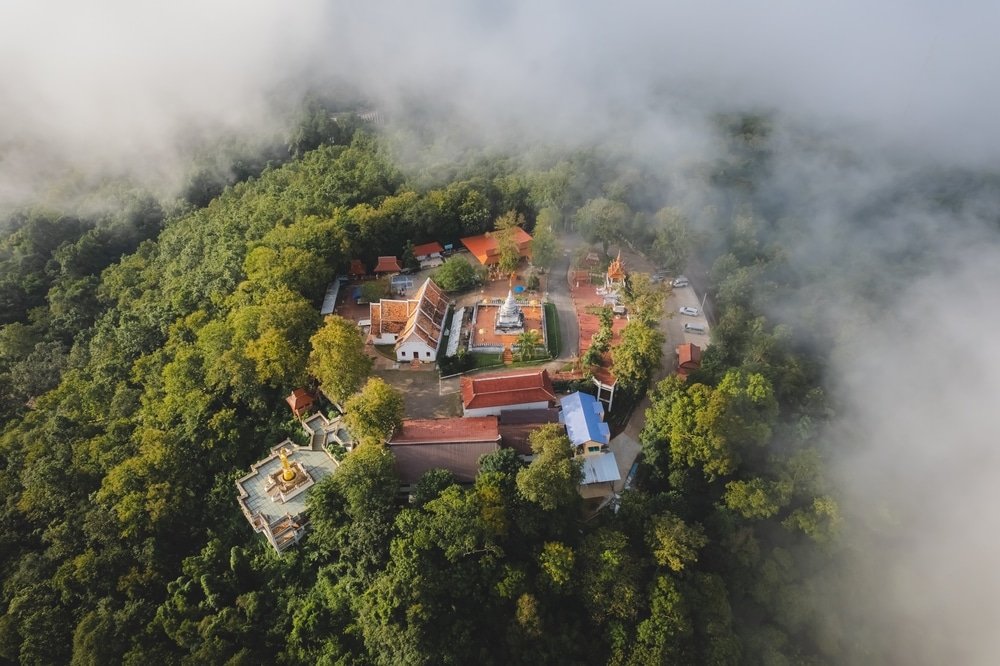 ภาพมุมสูงของกลุ่มวัดที่ล้อมรอบด้วยป่าเขียวชอุ่มหนาแน่นและบางส่วนปกคลุมด้วยหมอกหรือเมฆ แสดงให้เห็นหนึ่งในสถานที่ท่องเที่ยวที่สวยงามของน่านสำหรับผู้ที่ต้องการเที่ยวชมน่าน.
